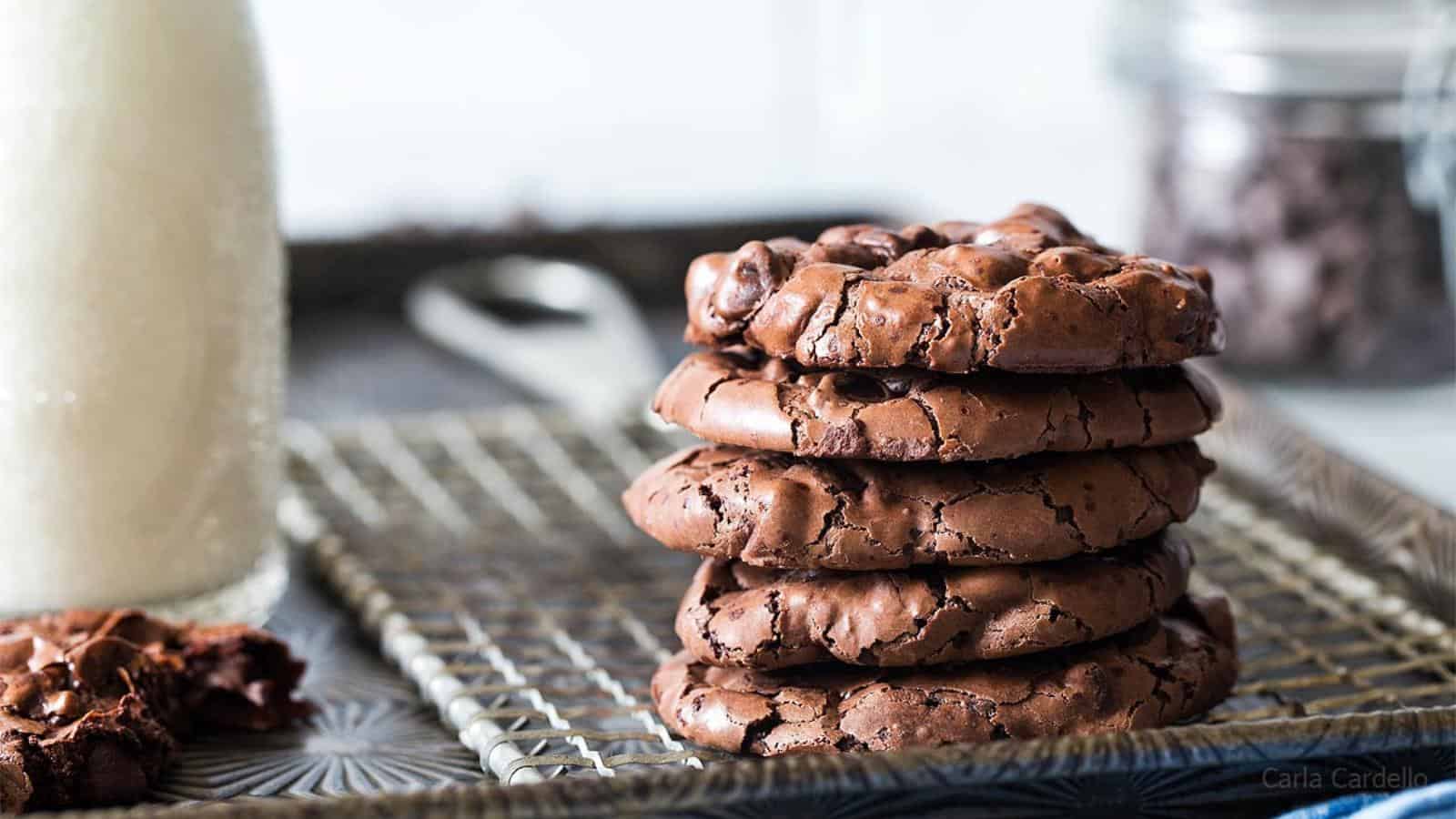 A stack of five chocolate cookies sits on a wire rack next to a glass bottle of milk and a jar of chocolate chips in the background.