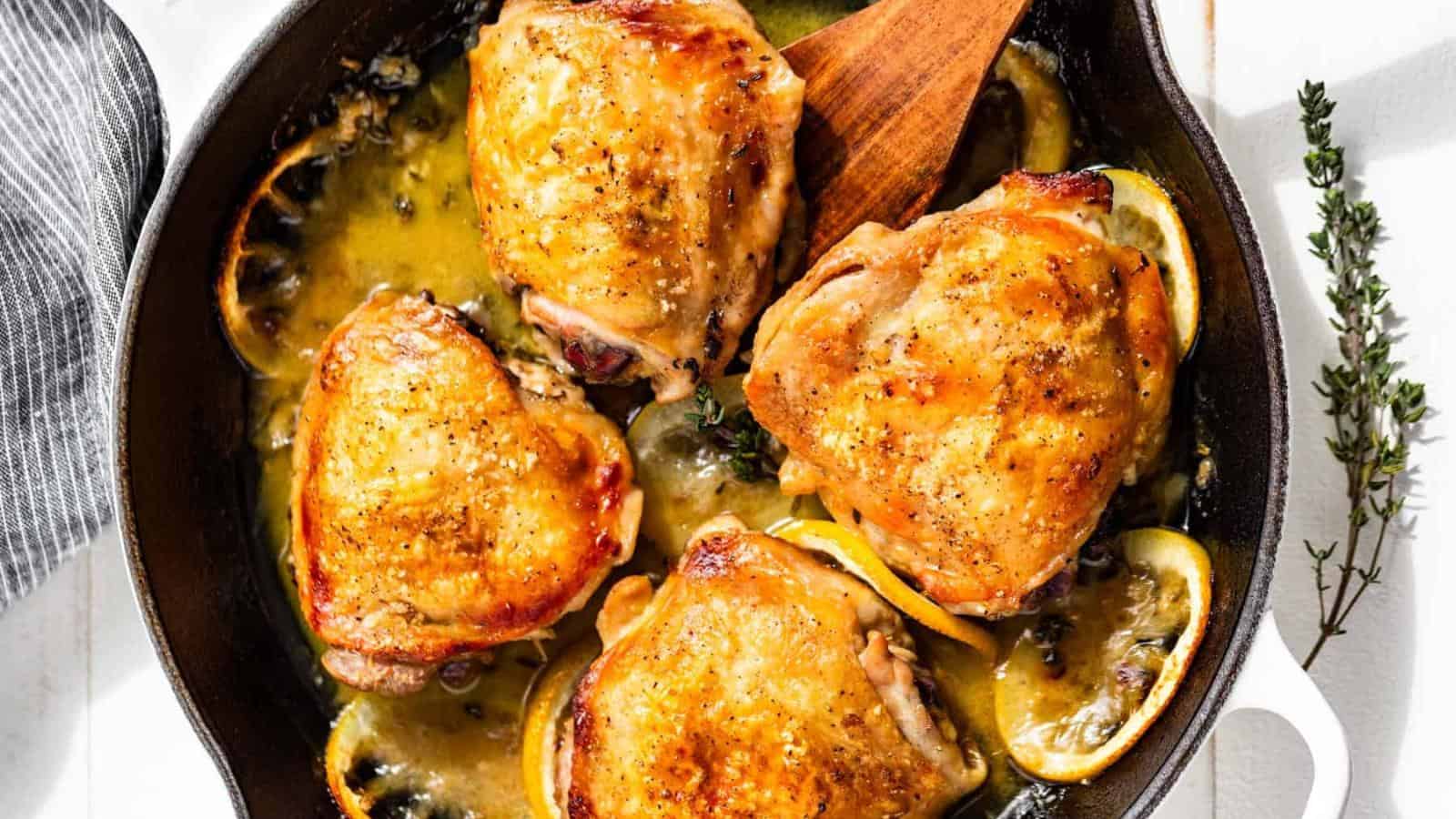 Four browned chicken thighs are cooked in a cast iron skillet with lemon slices and sauce, garnished with fresh thyme.
