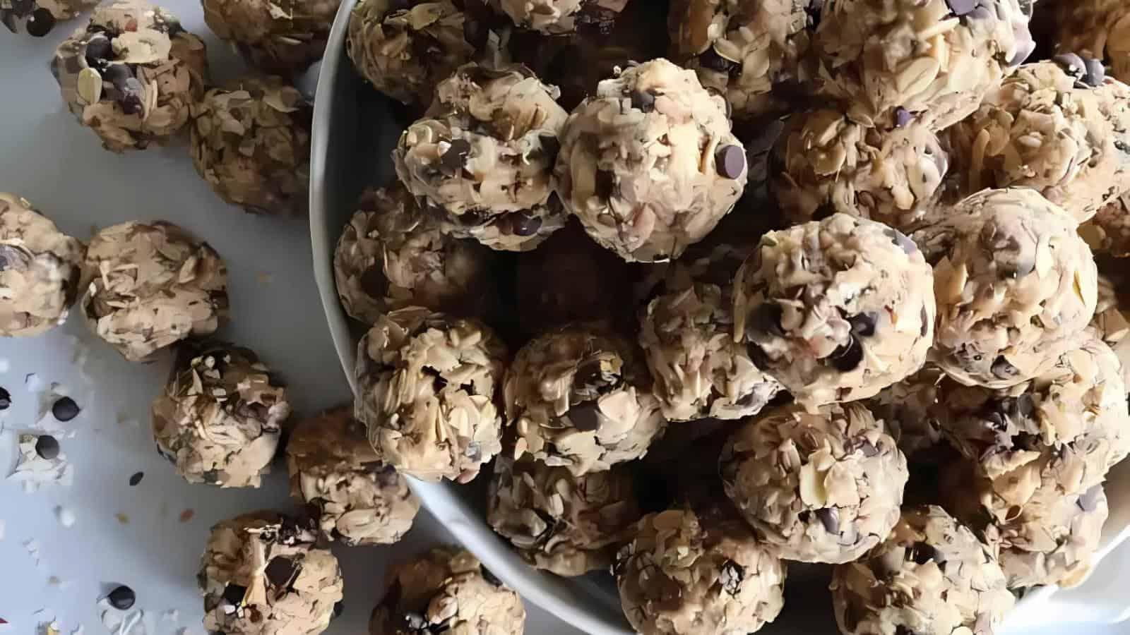 A white bowl filled with oat and chocolate chip energy bites, with several more energy bites scattered on the surface nearby.