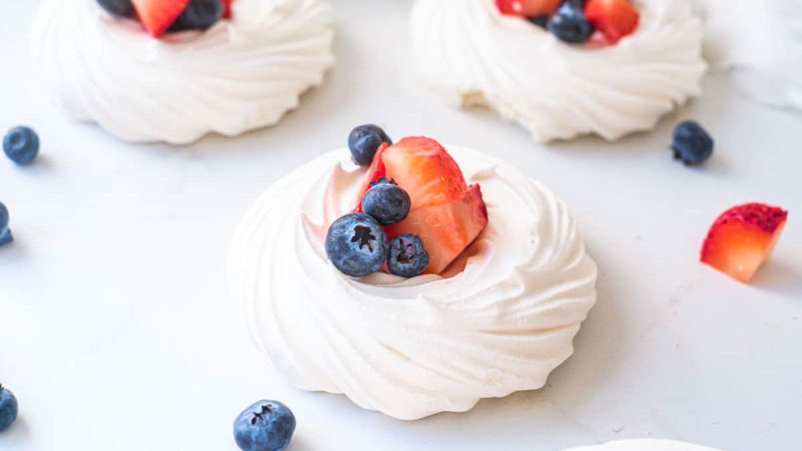 Mini pavlova desserts topped with sliced strawberries and blueberries, displayed on a white surface with scattered berries around them.