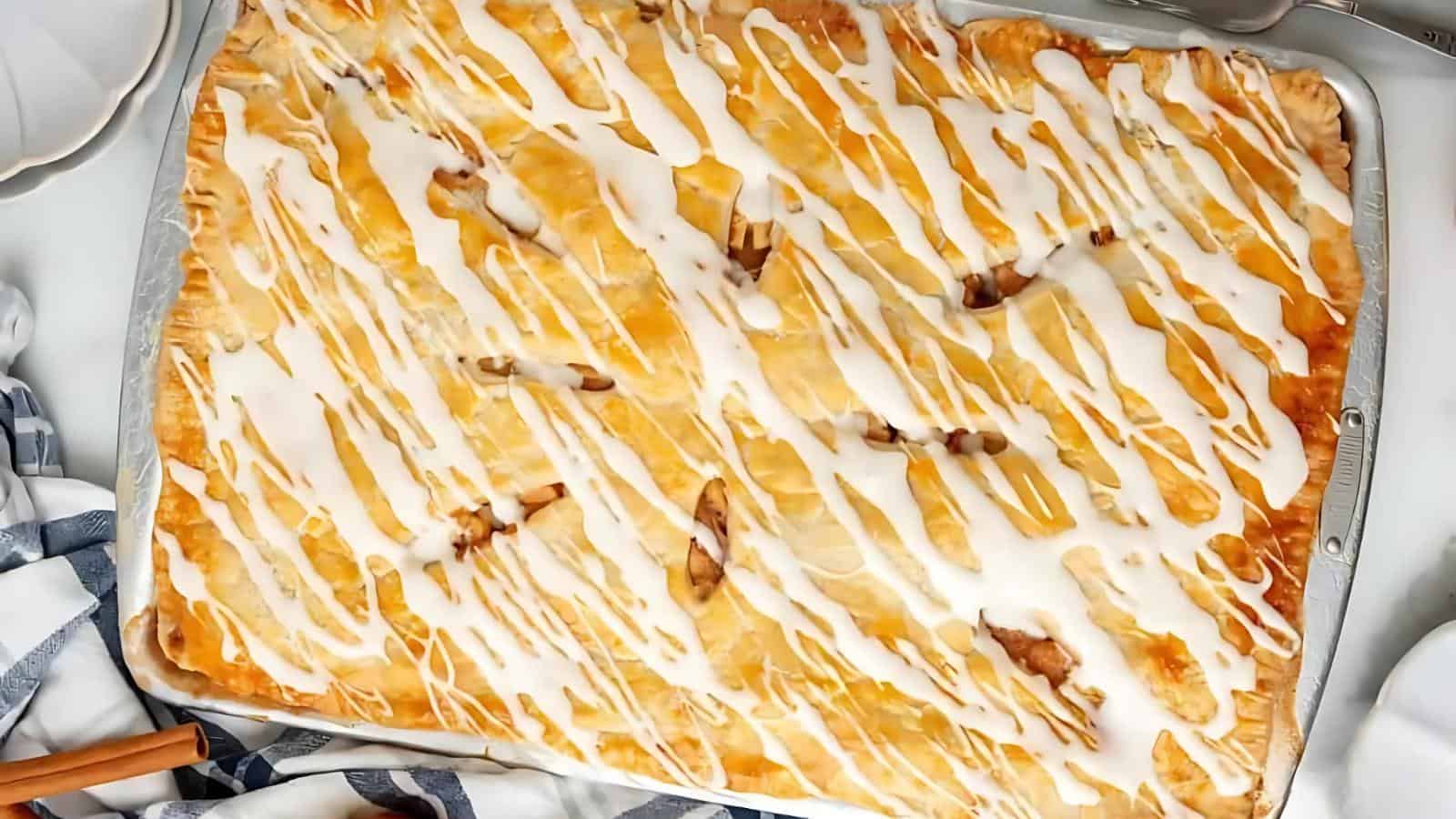 A large rectangular baked pastry with a golden-brown crust, topped with white icing drizzle, sits on a metal baking sheet.