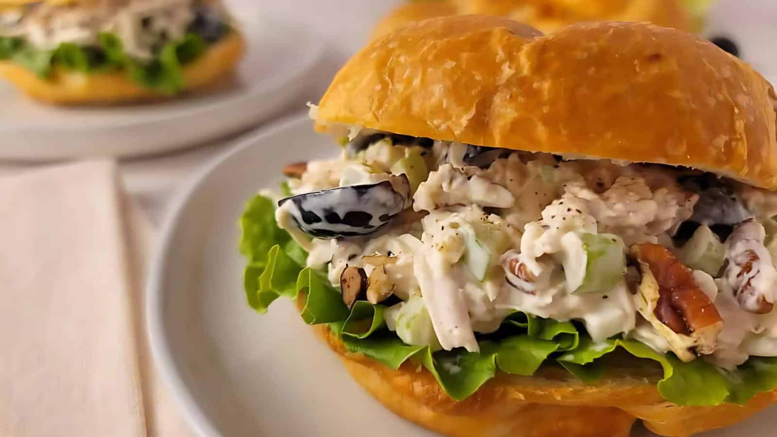 Close-up of a chicken salad sandwich with lettuce on a croissant, served on a white plate with another sandwich in the background.