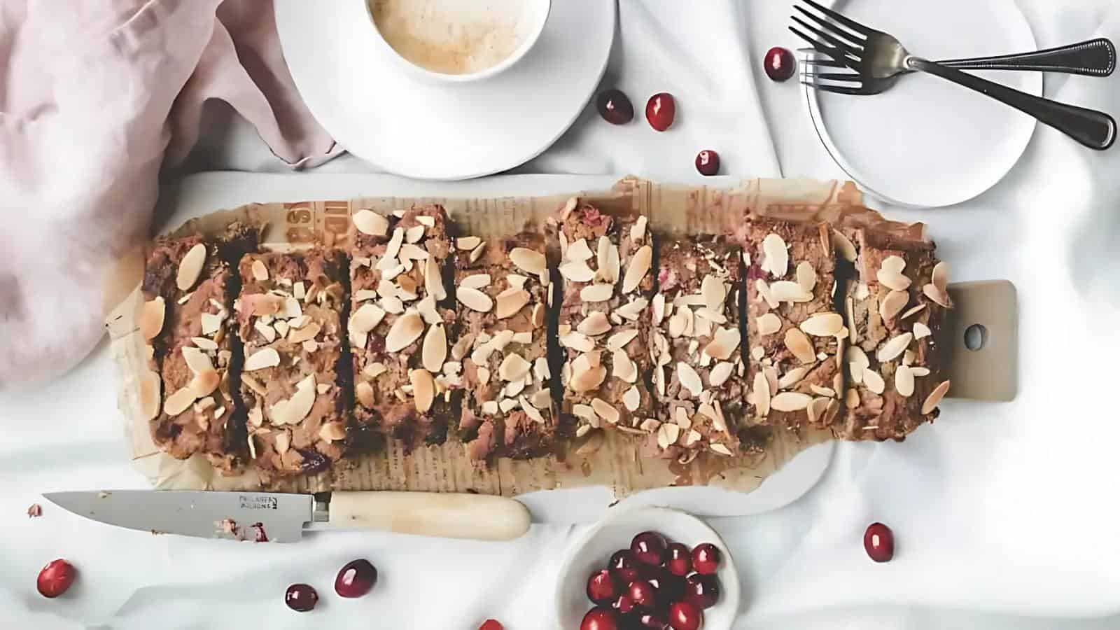 A sliced loaf topped with almond flakes is placed on parchment paper, with a knife, cranberries, cups, plates, and forks arranged nearby on a white surface.