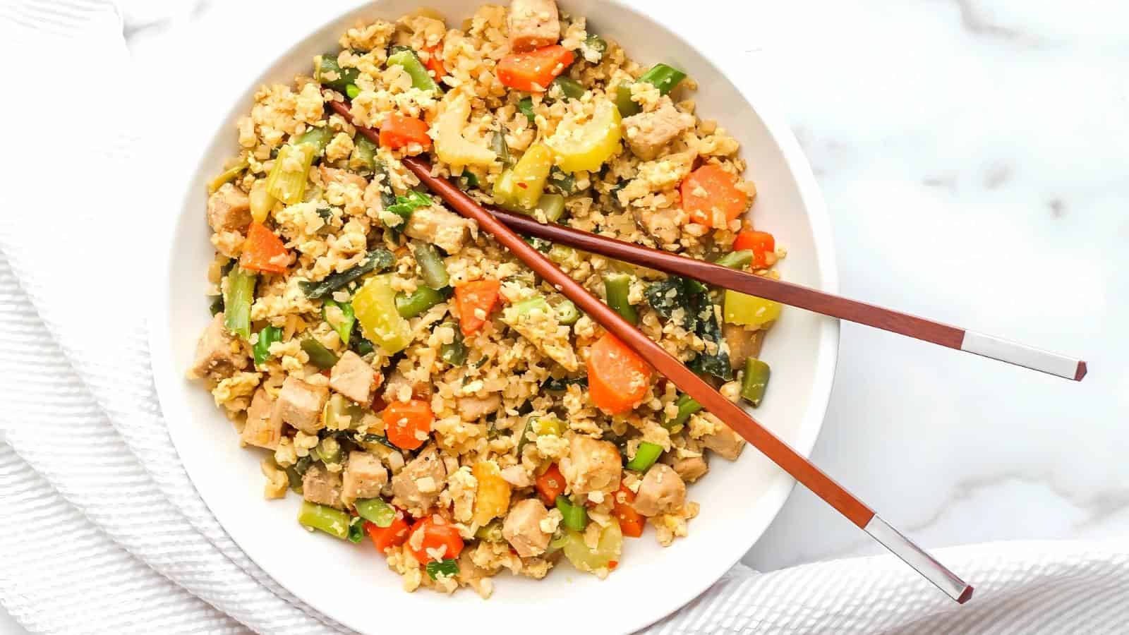 A bowl of stir-fried rice with diced chicken, scrambled eggs, carrots, green beans, and other vegetables, topped with a pair of brown chopsticks.