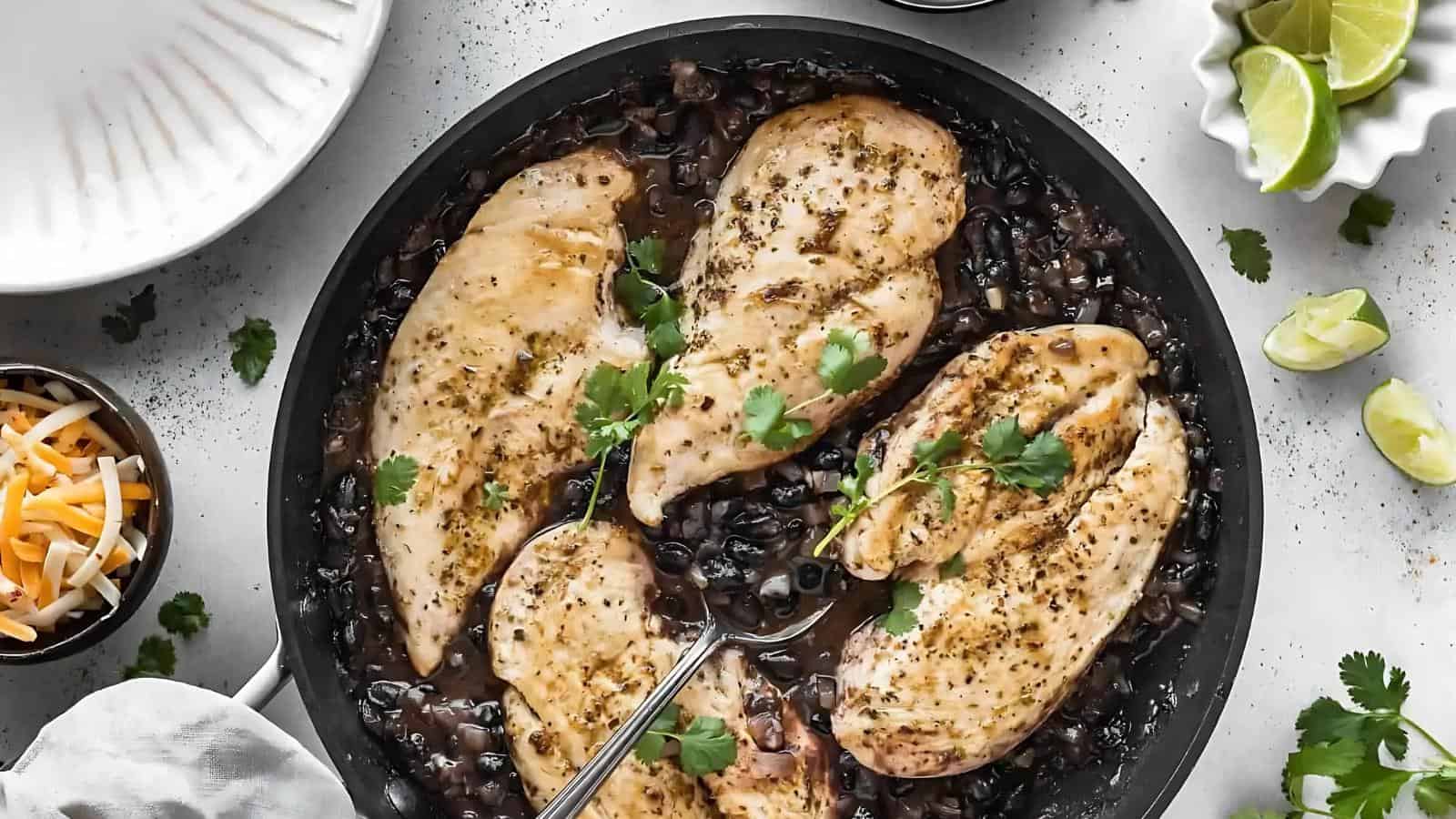 Four cooked chicken breasts garnished with cilantro rest on a bed of black beans and sauce in a black skillet, with a serving spoon placed in the pan.