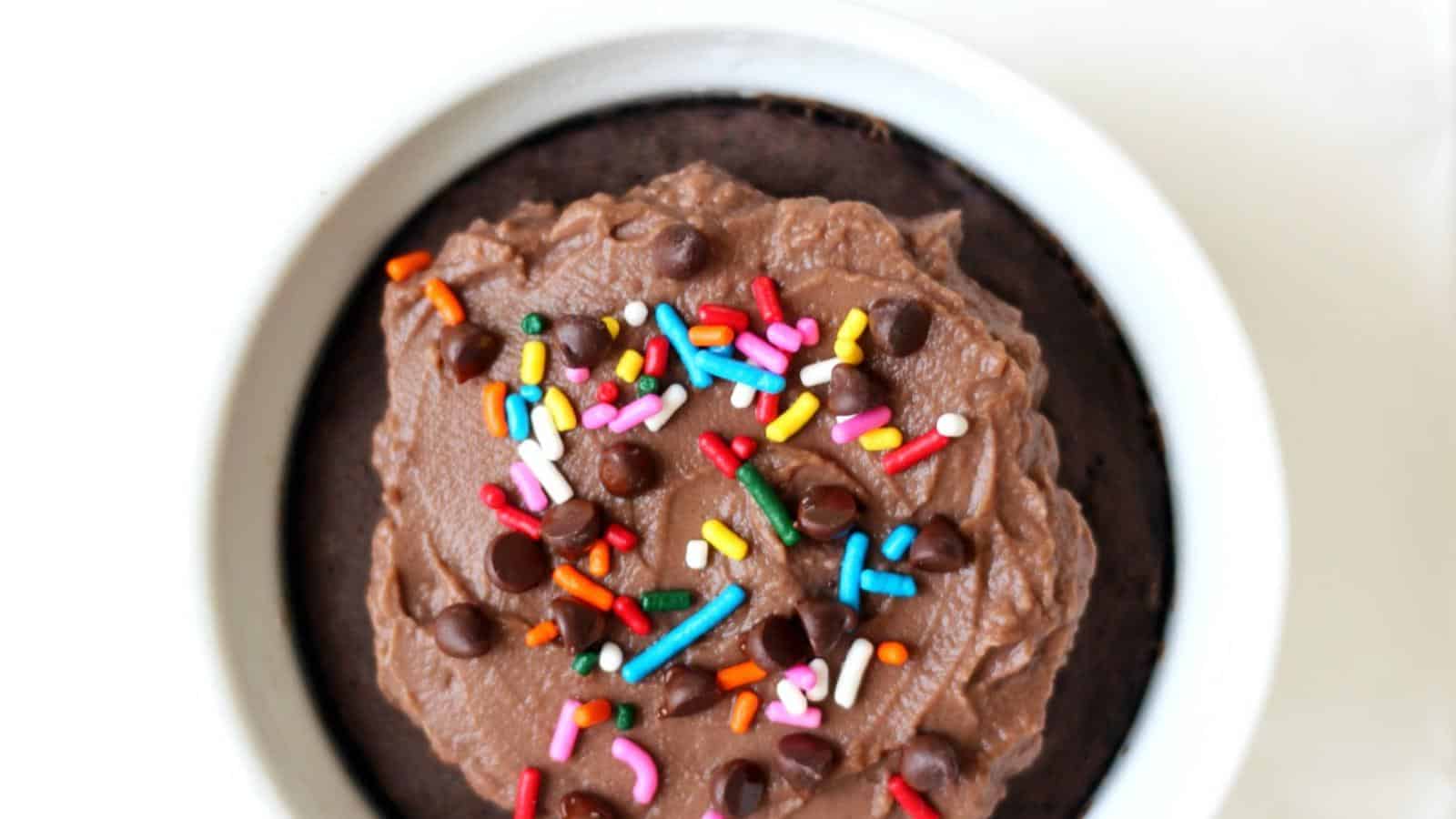 A chocolate dessert in a white bowl topped with chocolate frosting, rainbow sprinkles, and chocolate chips.