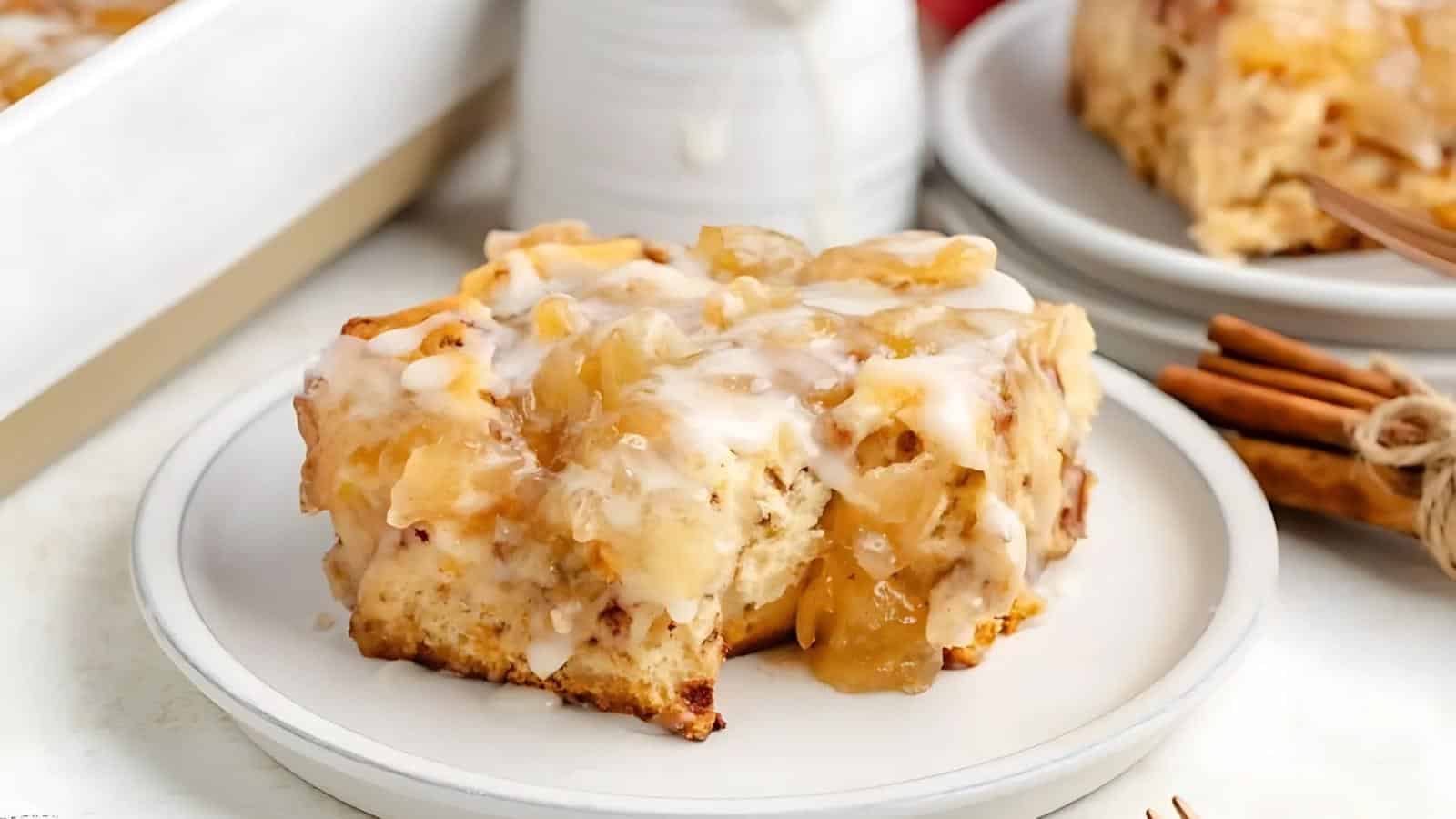 A piece of cinnamon roll topped with apple filling and white icing is served on a white plate, with a bundle of cinnamon sticks and another plate in the background.
