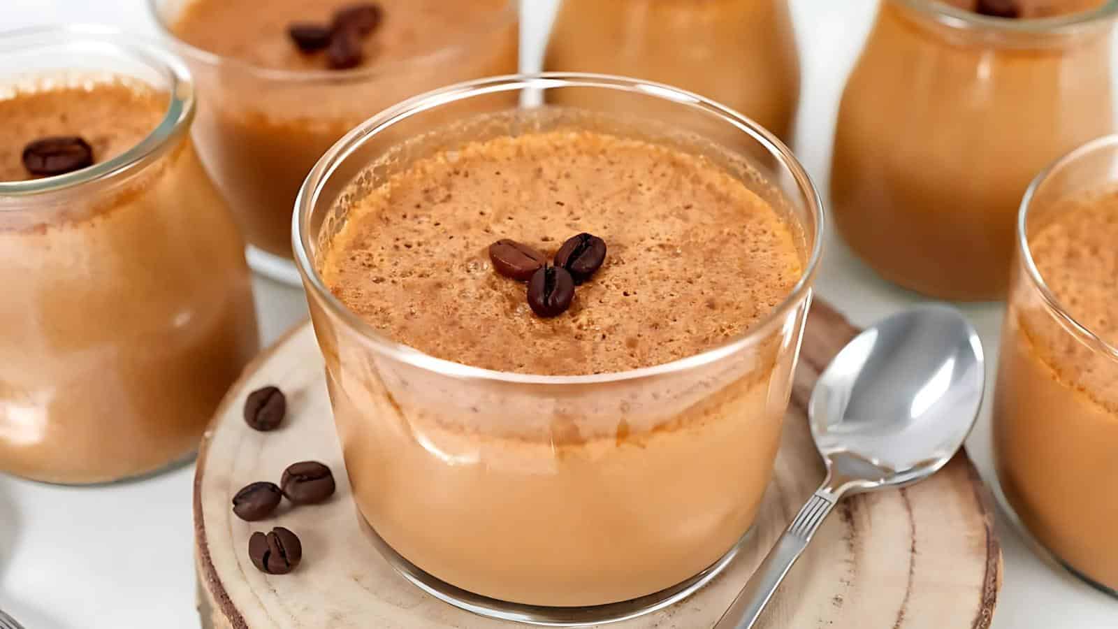 Glass jars filled with coffee-flavored mousse, each topped with coffee beans, sit on a wooden surface next to a silver spoon.