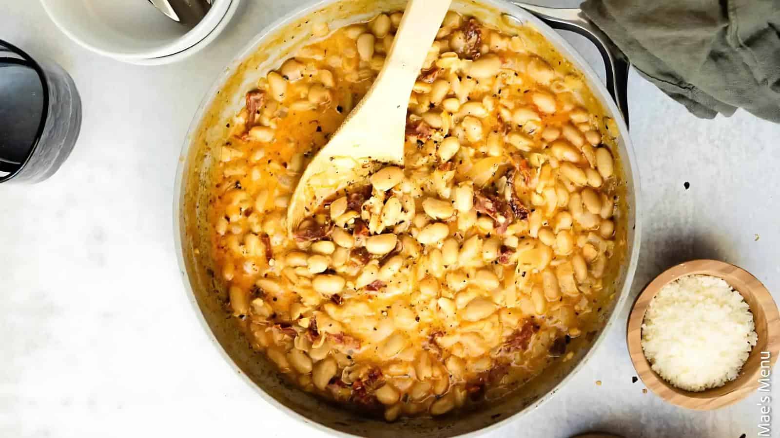A pot of creamy white beans with bacon and herbs is stirred with a wooden spoon; a small bowl of grated cheese sits nearby.