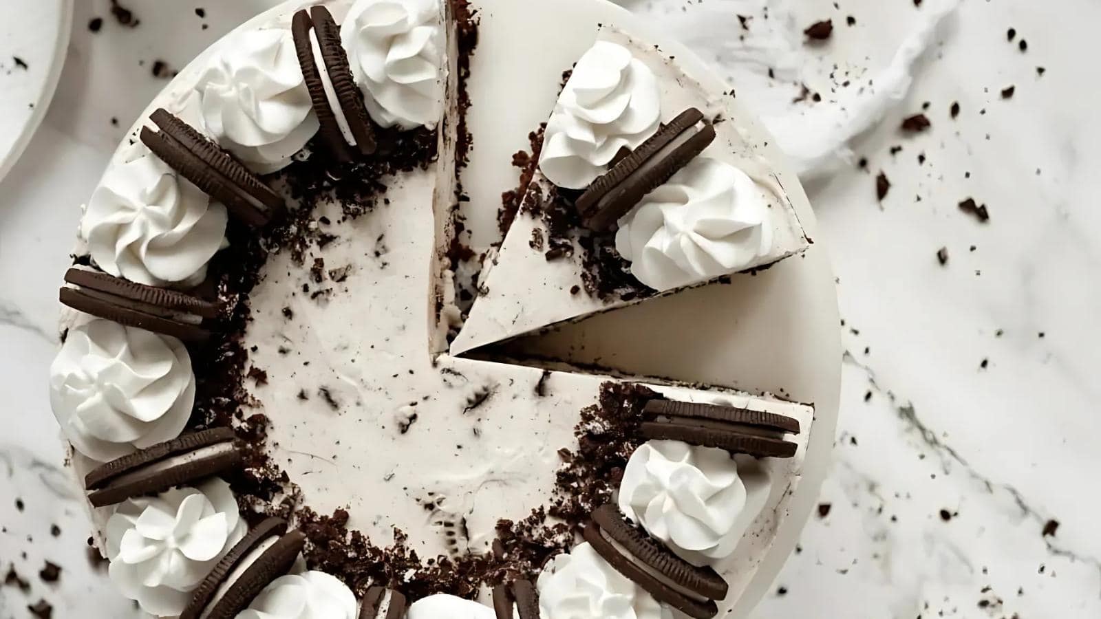 A cookies and cream cake with a single slice removed, topped with whipped cream swirls and chocolate sandwich cookies, on a white surface with scattered crumbs.