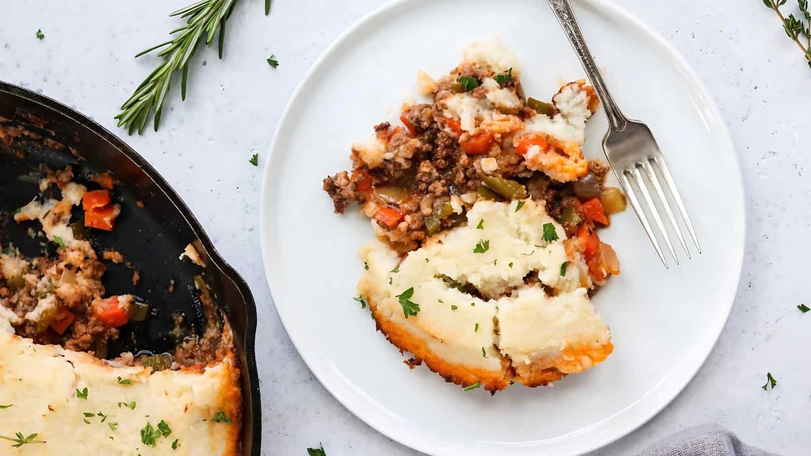 A serving of shepherd's pie with mashed potato topping, ground meat, and vegetables on a white plate, next to a skillet and garnished with herbs.