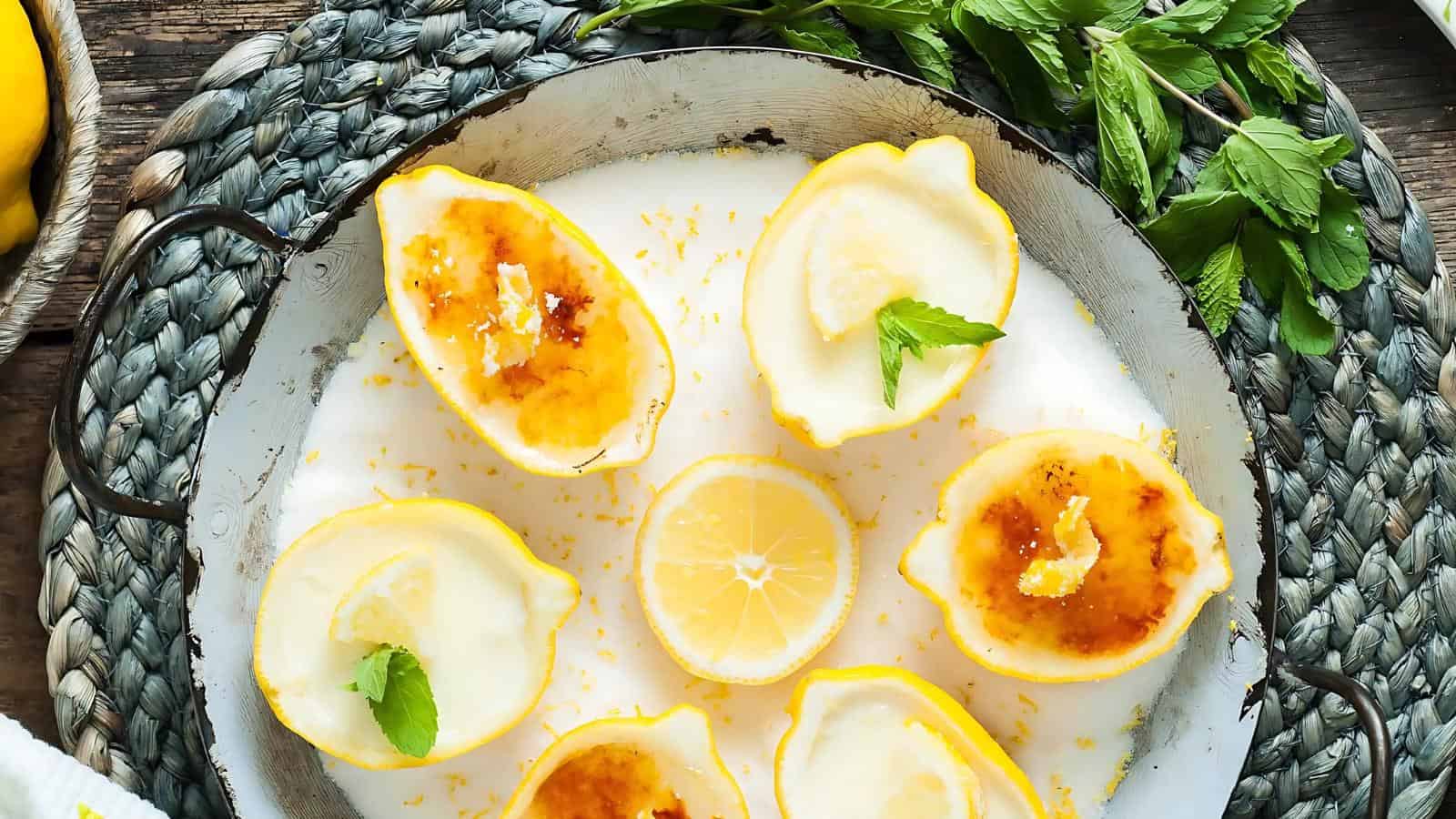 Halved lemons filled with a creamy dessert, some topped with caramelized sugar and mint leaves, arranged on a white plate with a sliced lemon and fresh mint nearby.