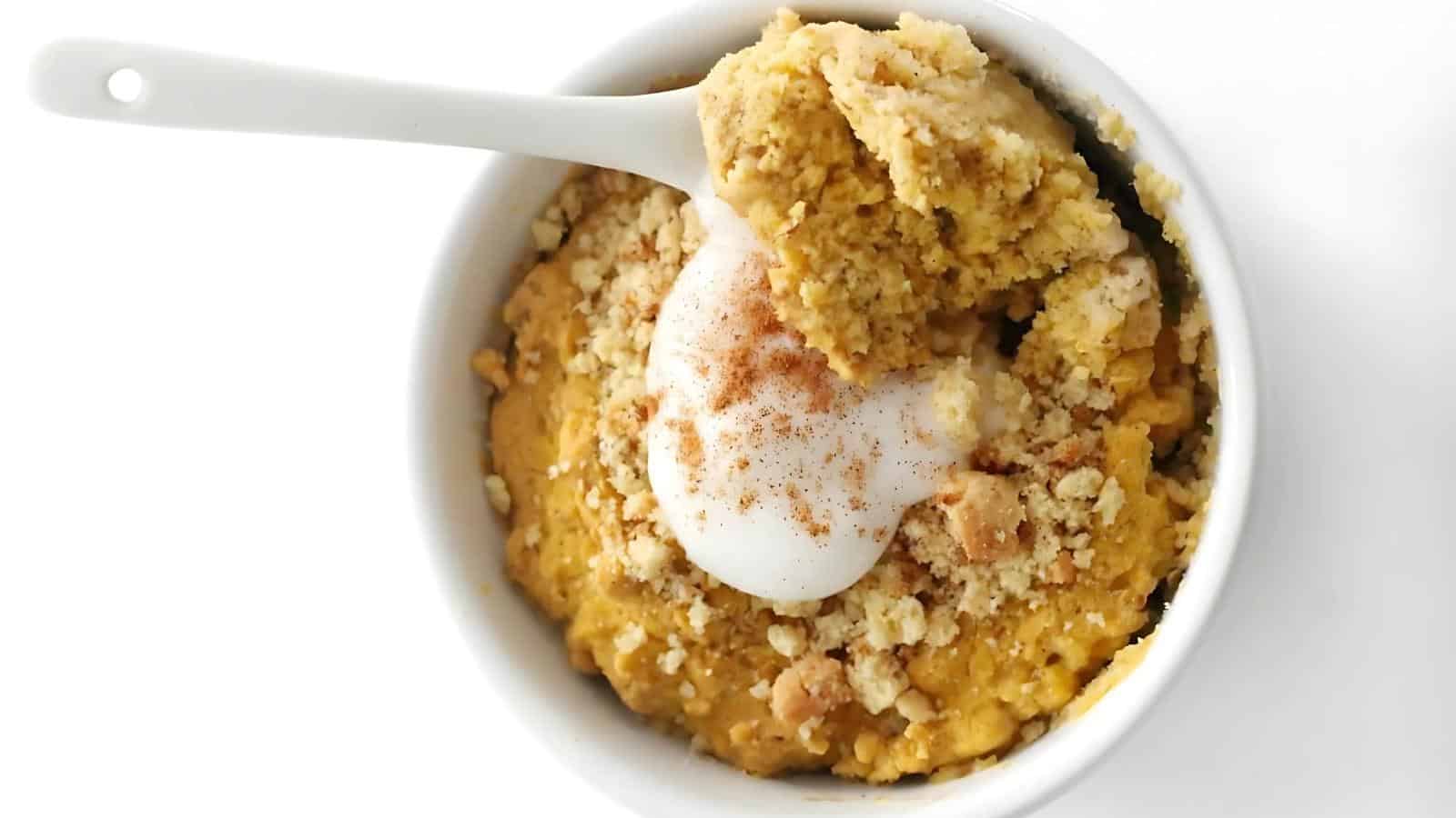 A white bowl filled with a crumbly, golden cake topped with a dollop of whipped cream and a sprinkle of cinnamon, with a spoon taking a bite.