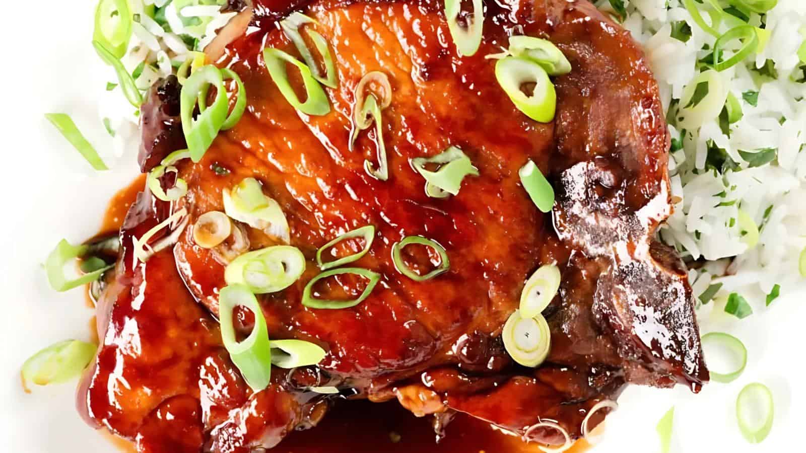 Glazed pork chops topped with sliced green onions, served next to a portion of white rice with herbs on a white plate.