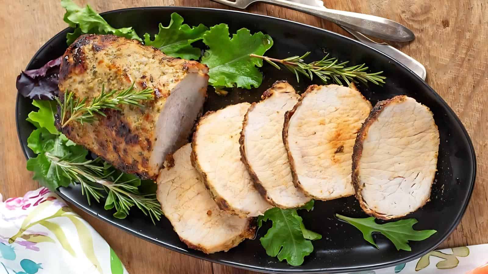 A sliced roasted pork loin garnished with fresh rosemary and leafy greens is arranged on a black oval platter with serving utensils beside it.