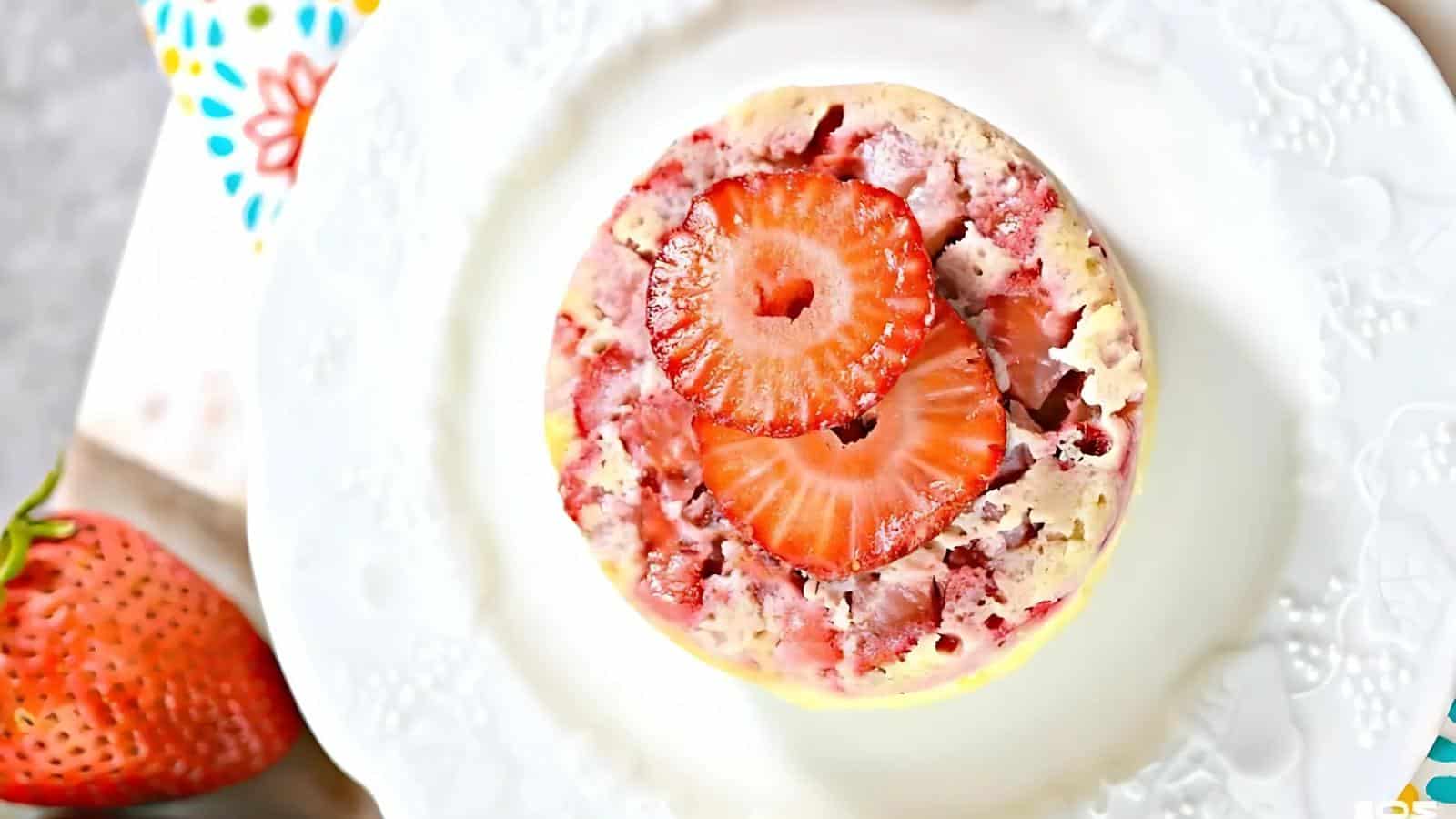 A round strawberry mug cake topped with two strawberry slices sits on a white decorative plate, with a whole strawberry nearby.