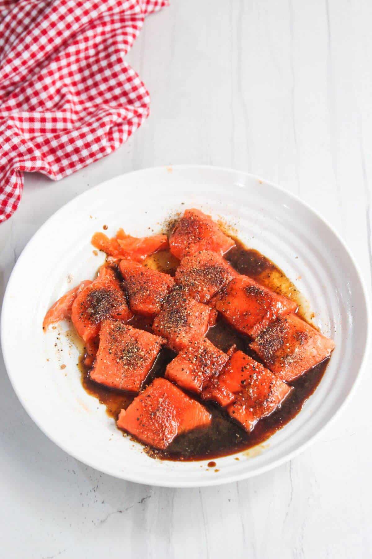 A white plate contains pieces of raw salmon in a dark marinade, topped with ground black pepper. A red and white checkered cloth is nearby.