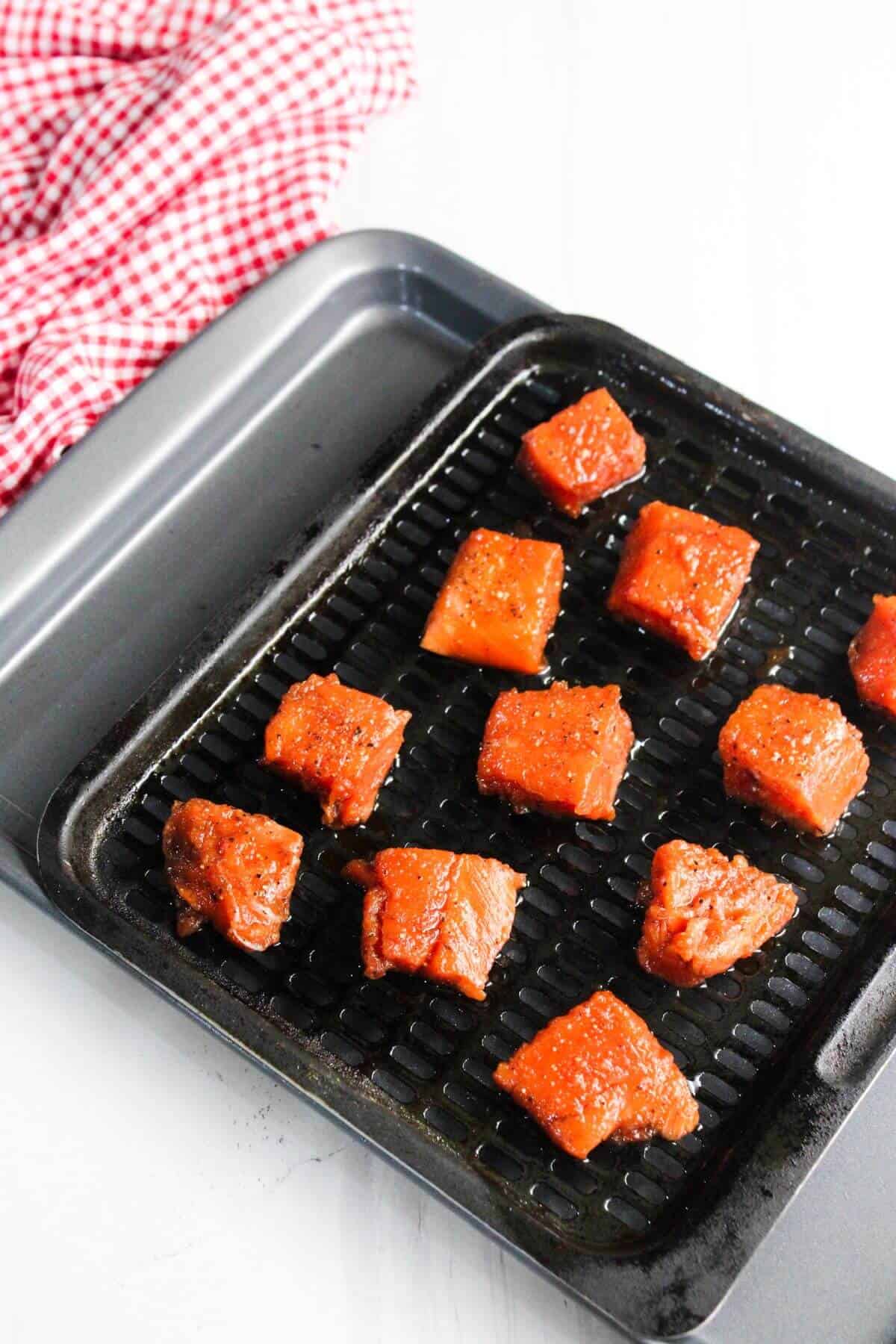 Cubed pieces of seasoned salmon are arranged on a black grill tray over a baking sheet, with a red and white checkered cloth in the background.