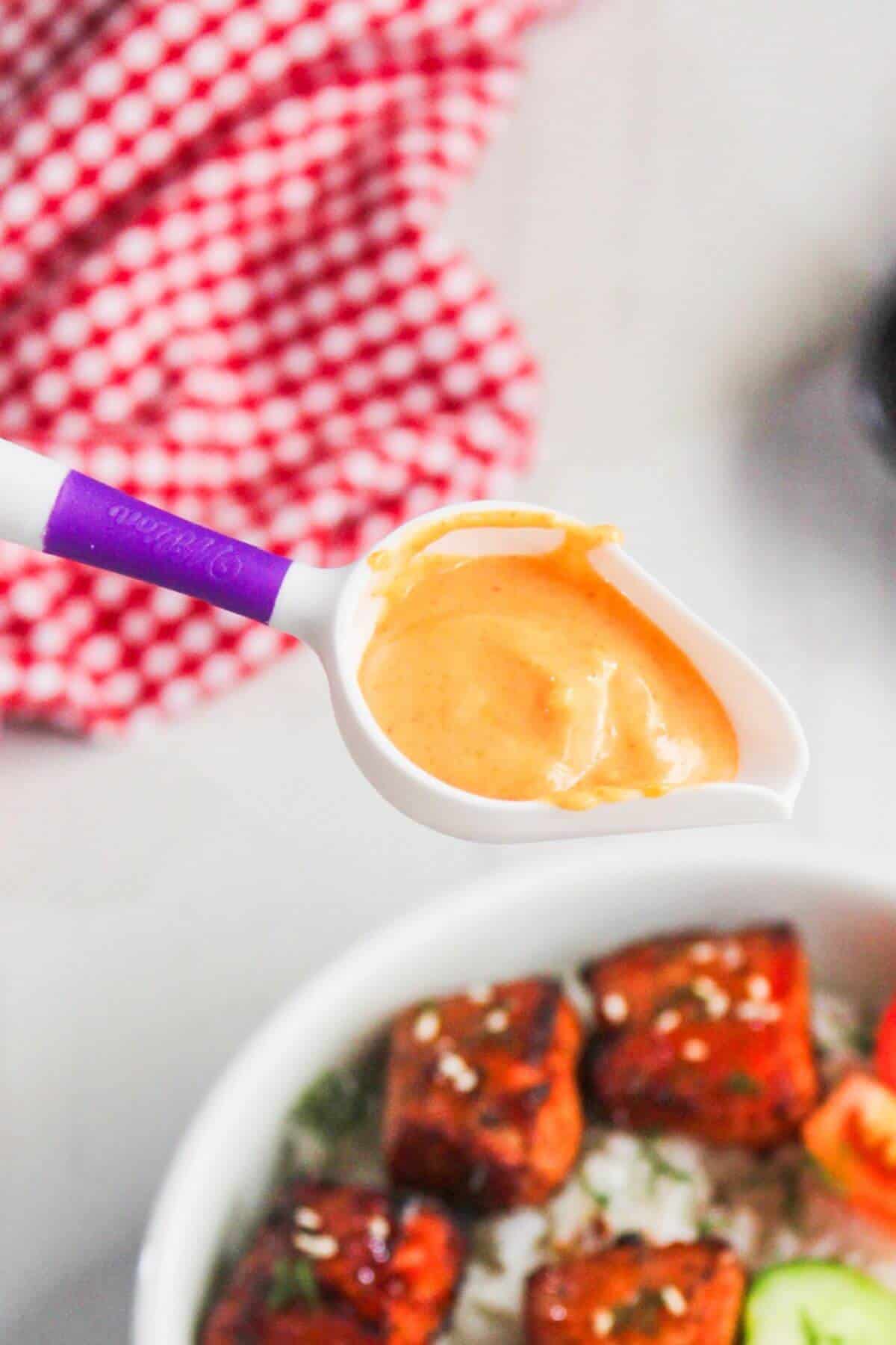 A white and purple measuring spoon holds a creamy orange sauce above a bowl of rice topped with glazed tofu cubes and sliced cucumbers.
