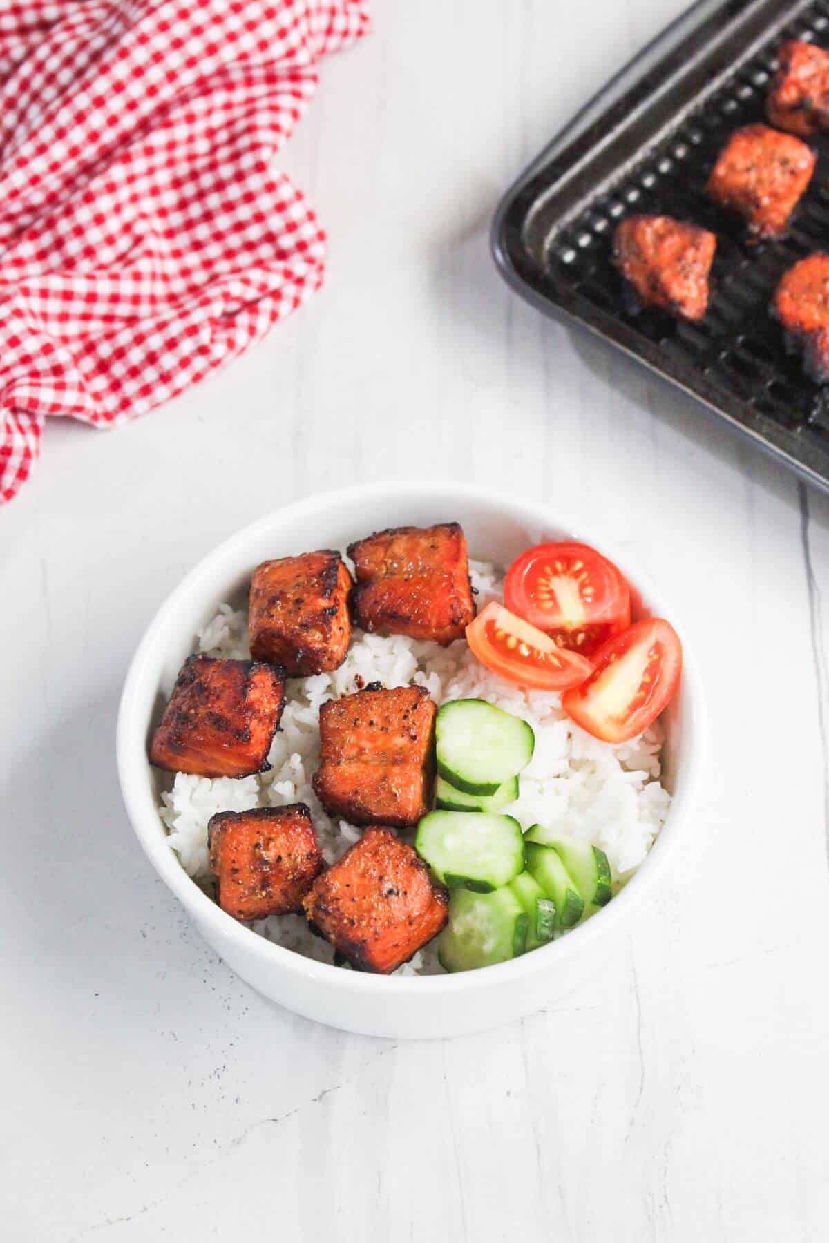 A bowl of white rice topped with grilled tofu cubes, sliced cucumber, and halved cherry tomatoes. A tray with more tofu and a red checkered cloth are nearby.