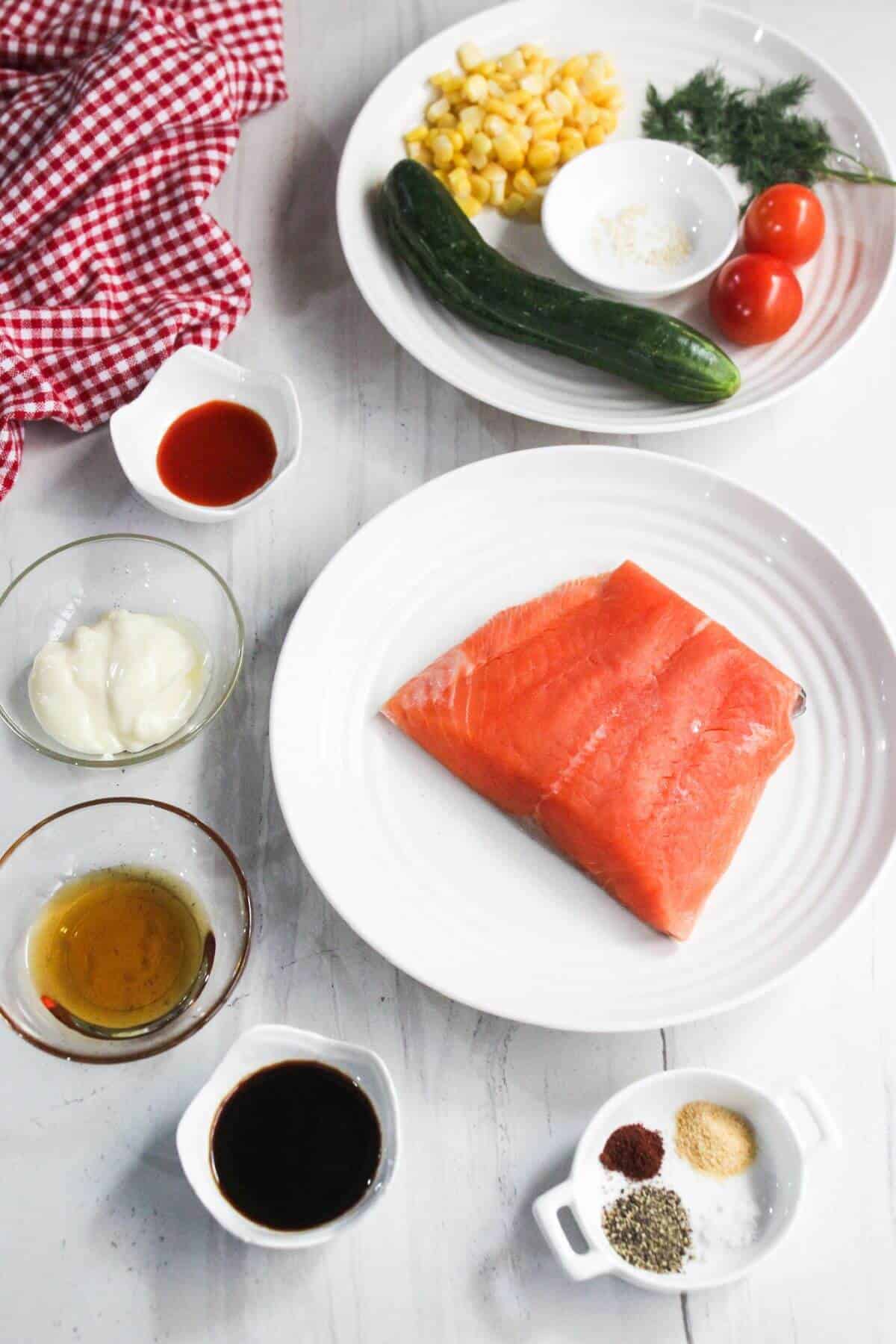 A raw salmon fillet on a plate surrounded by cucumber, cherry tomatoes, corn, mayonnaise, soy sauce, oil, seasoning, and a small bowl of hot sauce.