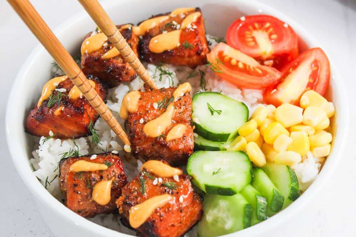 A bowl of white rice topped with glazed salmon cubes, sliced cucumber, corn, tomato wedges, and a drizzle of orange sauce, with chopsticks picking up a piece.