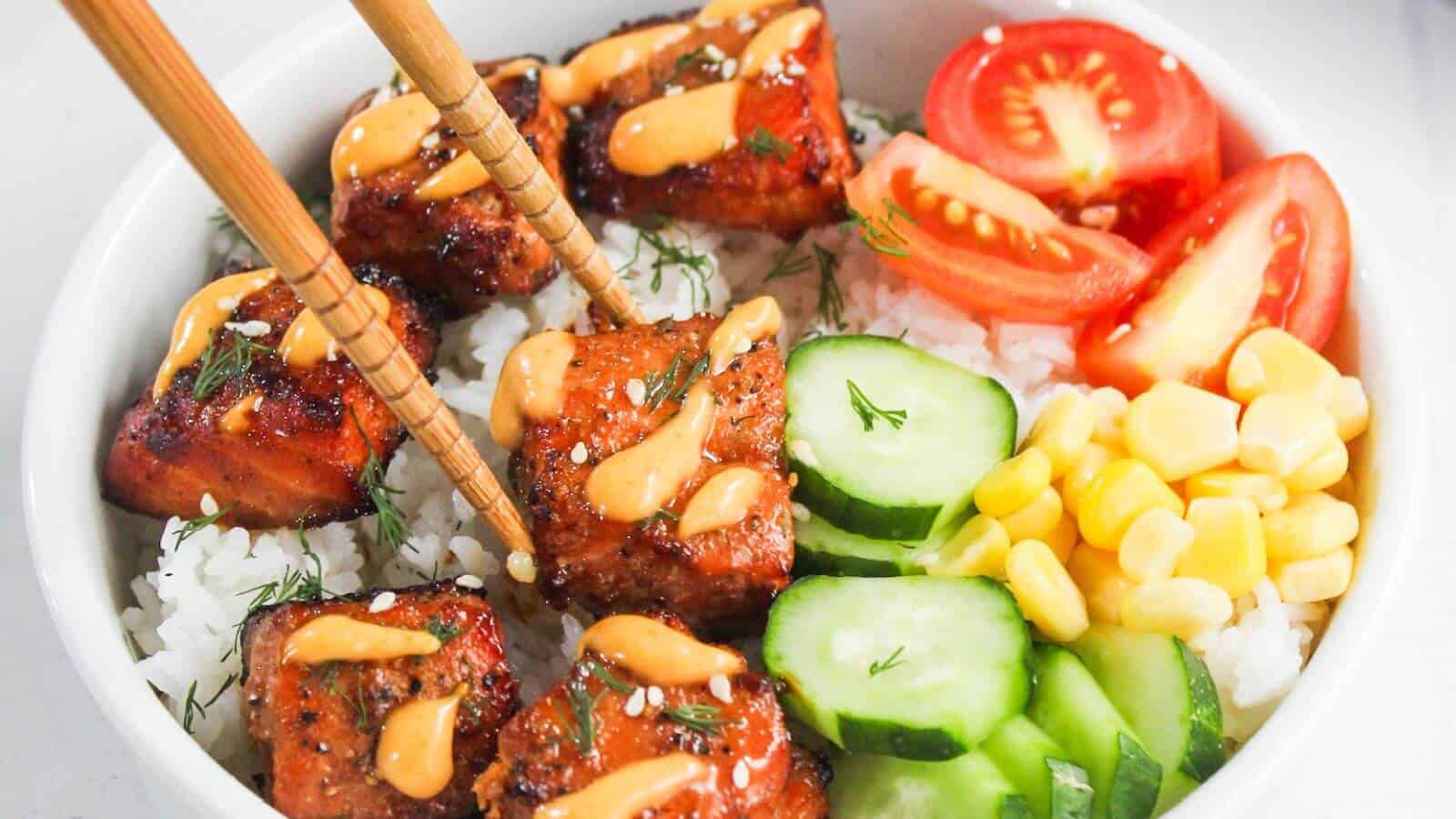A bowl of white rice topped with glazed salmon, sliced cucumbers, corn, tomato wedges, and a drizzle of orange sauce, with chopsticks picking up a piece of salmon.