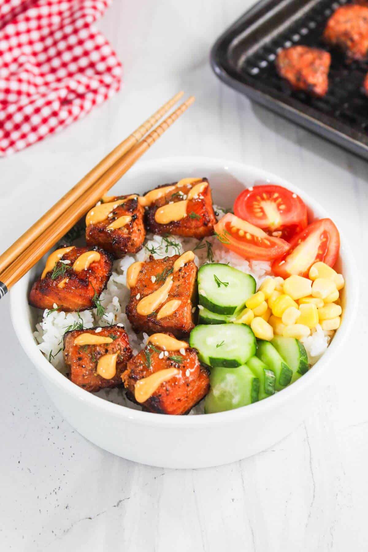 A white bowl filled with rice, glazed tofu cubes, sliced cucumber, corn, and tomato, topped with sauce. Chopsticks rest on the bowl's edge.
