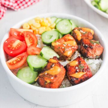 A white bowl filled with rice, grilled salmon pieces, cucumber slices, cherry tomatoes, corn, and a drizzle of sauce.