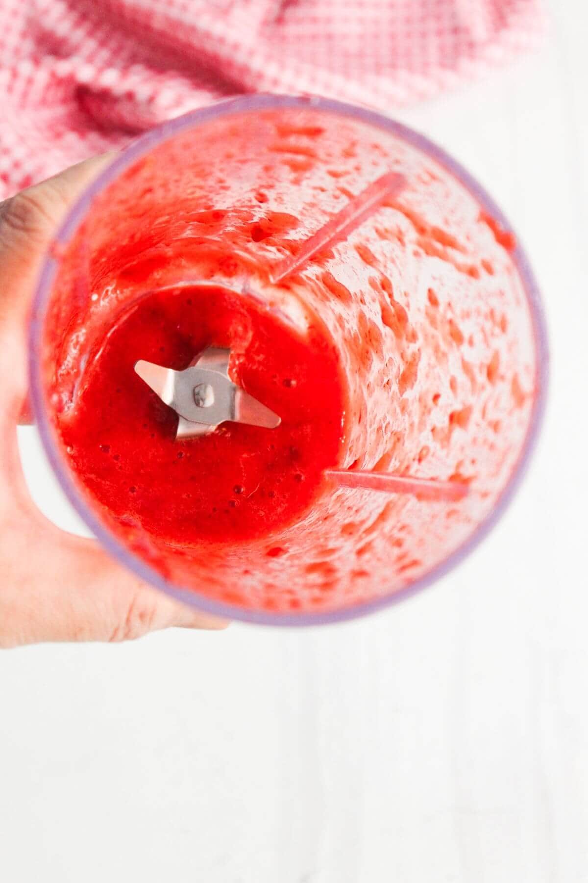 A hand holds a blender cup containing a bright red, blended fruit mixture with visible blades at the bottom. A pink cloth is in the background.