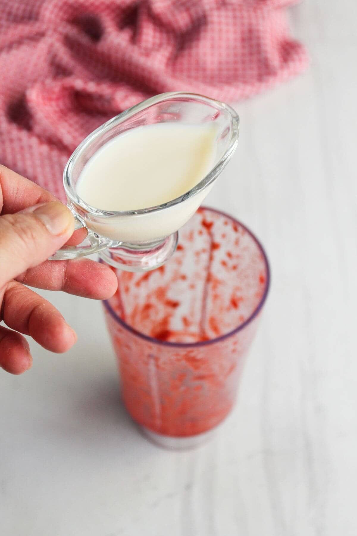 A hand holds a small glass pitcher of cream above a blender cup containing a red fruit mixture, with a pink cloth in the background.