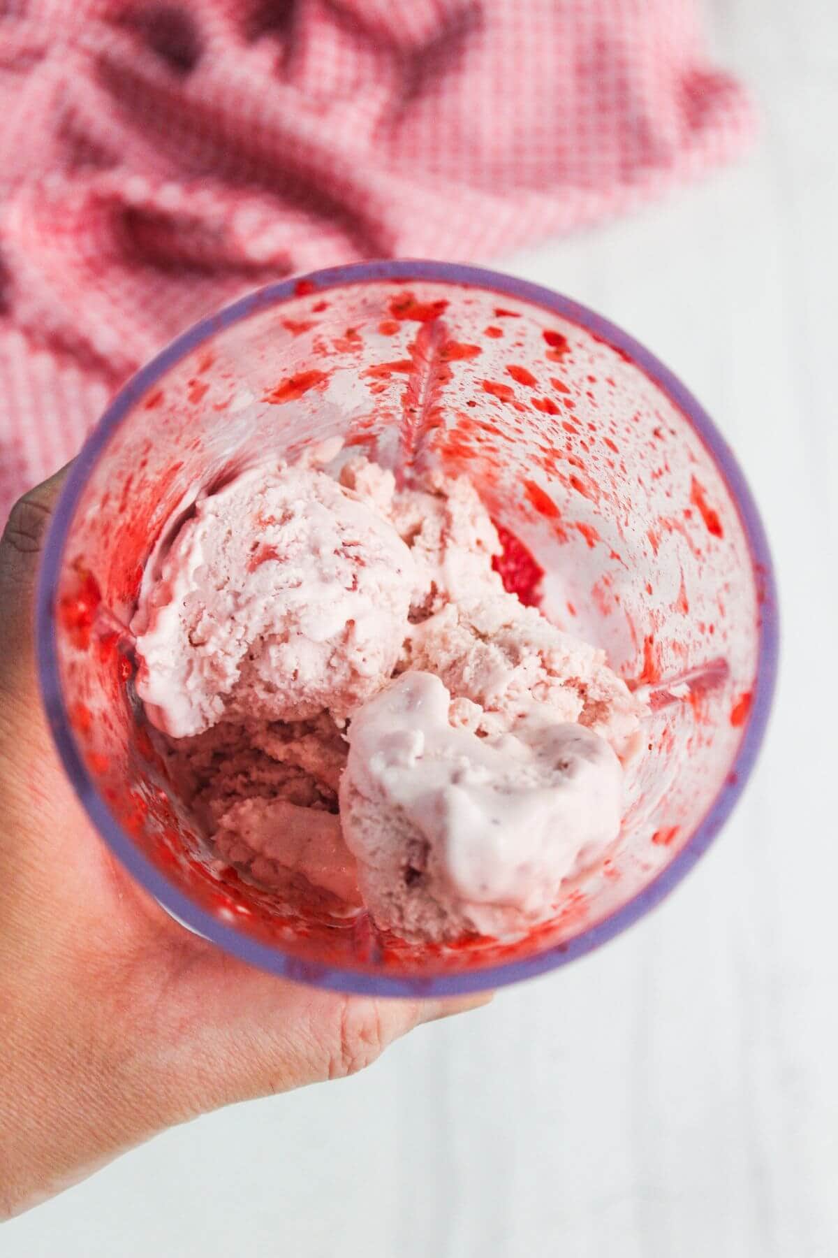 A hand holds a transparent cup containing pink ice cream with red sauce smeared on the sides, with a pink cloth in the blurred background.
