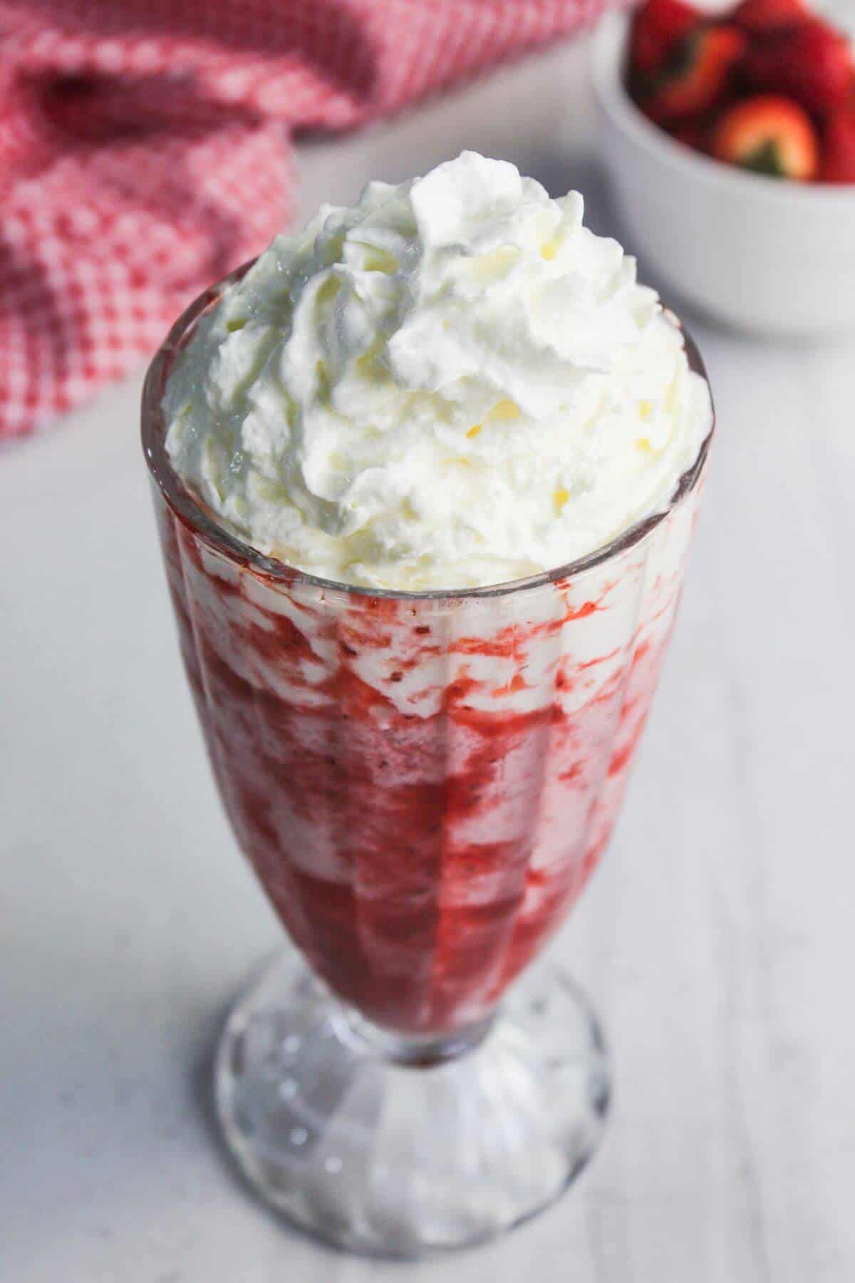 A glass filled with a strawberry milkshake, topped with whipped cream, sits on a white surface with a bowl of strawberries and a red cloth in the background.