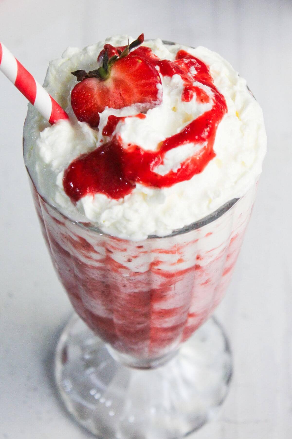 A glass filled with a strawberry milkshake topped with whipped cream, strawberry sauce, a whole strawberry, and a red-striped straw.