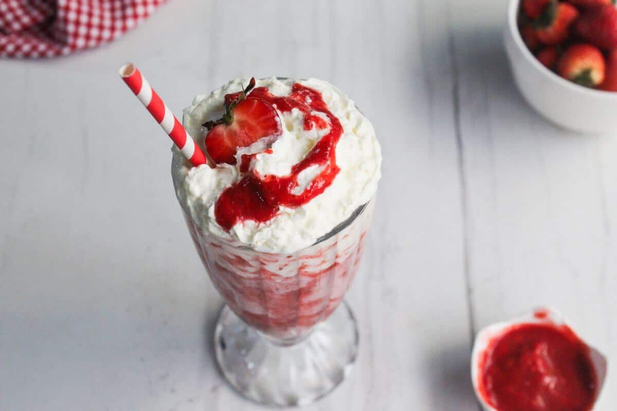 A strawberry milkshake topped with whipped cream, strawberry sauce, a fresh strawberry, and a red striped straw, on a white table with bowls of strawberries and sauce in the background.