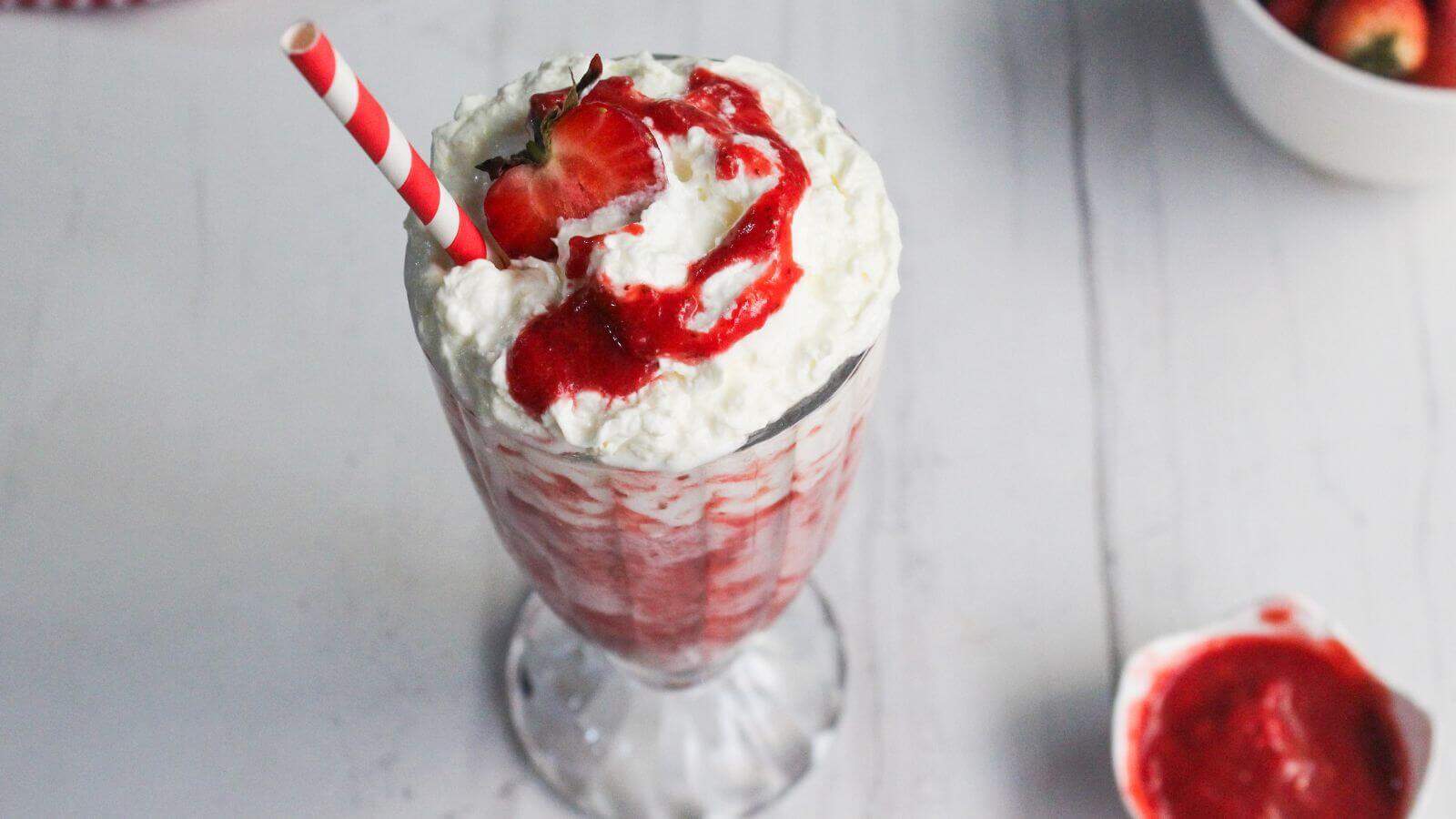 A strawberry milkshake topped with whipped cream, strawberry sauce, a fresh strawberry, and a red striped straw, on a white table with bowls of strawberries and sauce in the background.