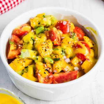 A white bowl filled with a colorful salad of chopped tomatoes, avocado, and greens, topped with a creamy yellow dressing and sprinkled with black pepper.