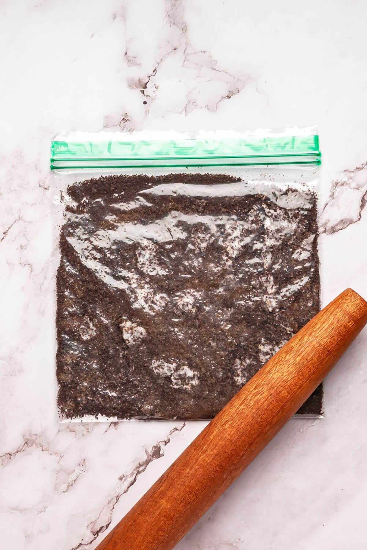 A plastic ziplock bag filled with crushed dark crumbs is on a marble surface next to a wooden rolling pin.
