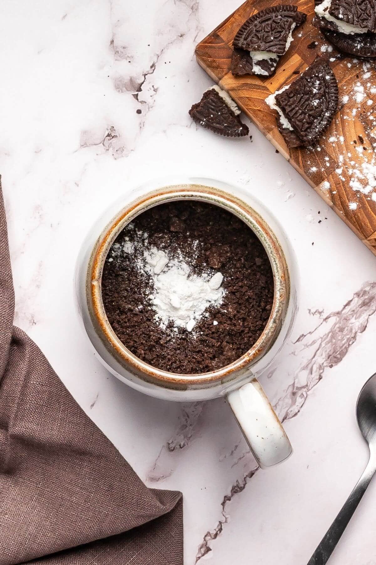 A mug filled with crushed chocolate cookies and white powder sits on a marble surface next to a wooden board with broken cookies and a brown cloth.