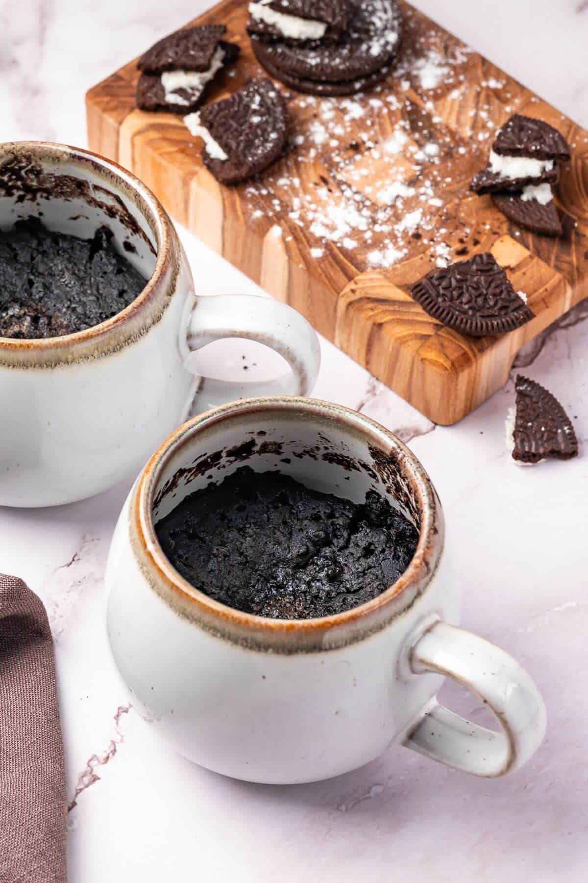 Two white mugs with chocolate mug cake sit on a marble surface, next to a wooden board with broken chocolate sandwich cookies and crumbs.