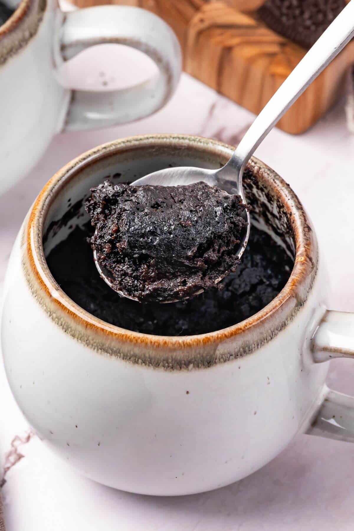 A spoon lifts a portion of chocolate mug cake from a white ceramic mug on a marble surface.