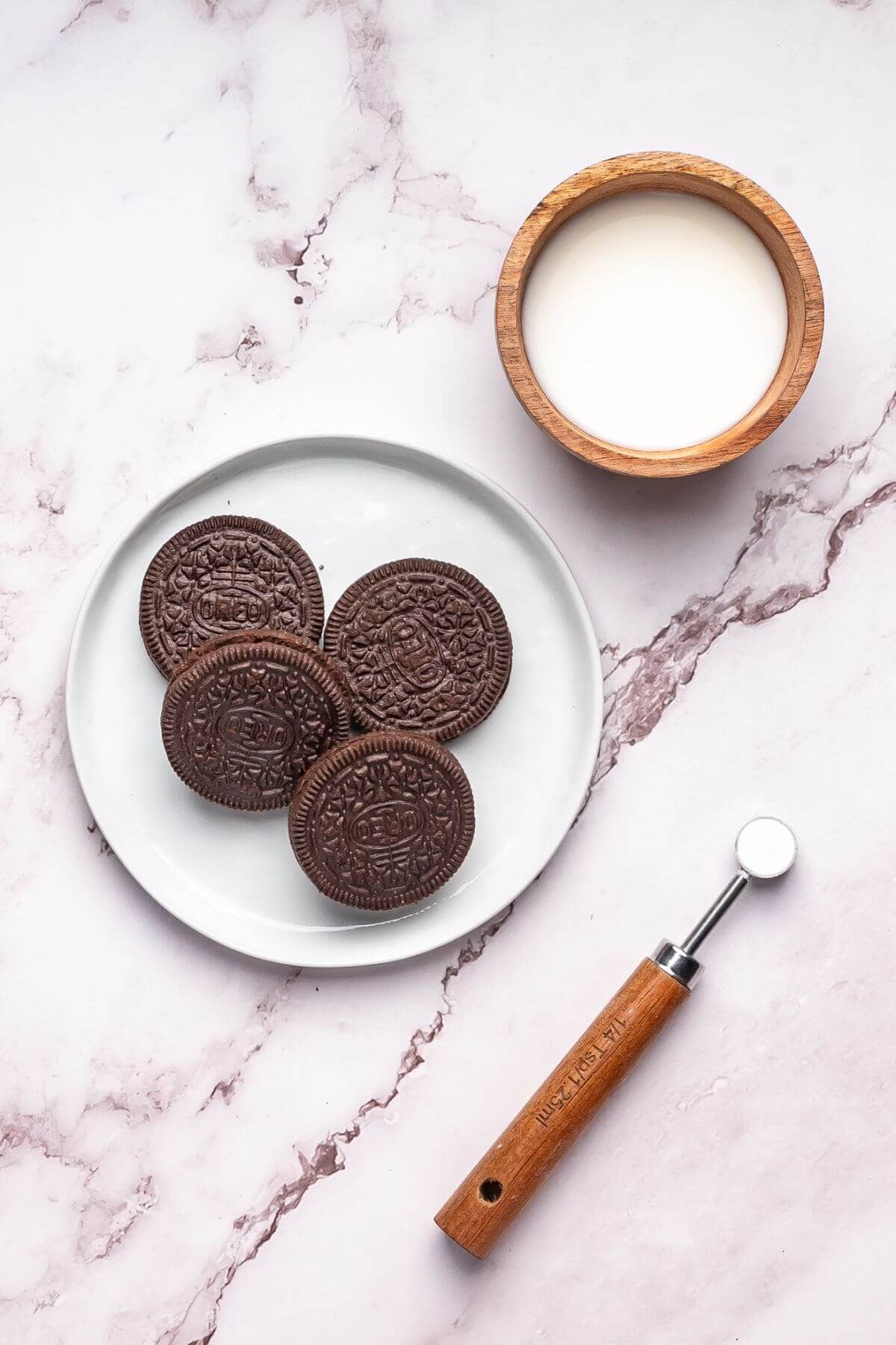 A plate with five chocolate sandwich cookies, a wooden bowl of milk, and a small metal and wood kitchen tool on a marble surface.