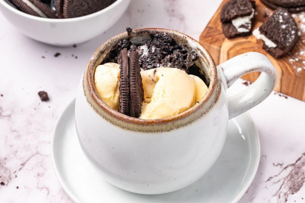 A white mug filled with crumbled chocolate cookies, vanilla ice cream, and a whole sandwich cookie, served on a white saucer.