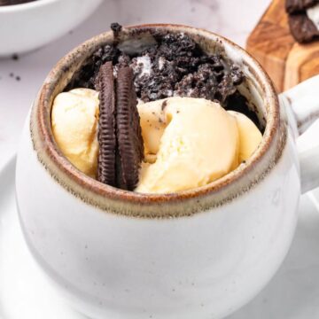 A white mug filled with vanilla ice cream, crushed chocolate sandwich cookies, and a whole cookie, placed on a saucer.