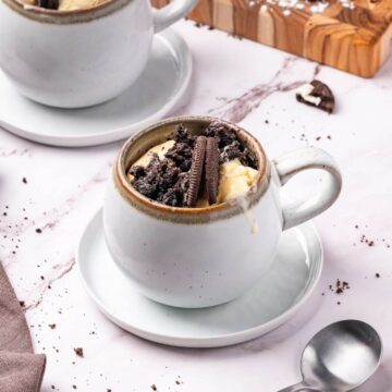 A white mug on a saucer filled with a dessert topped with crushed chocolate cookies and cream, placed on a marble surface with a spoon nearby.