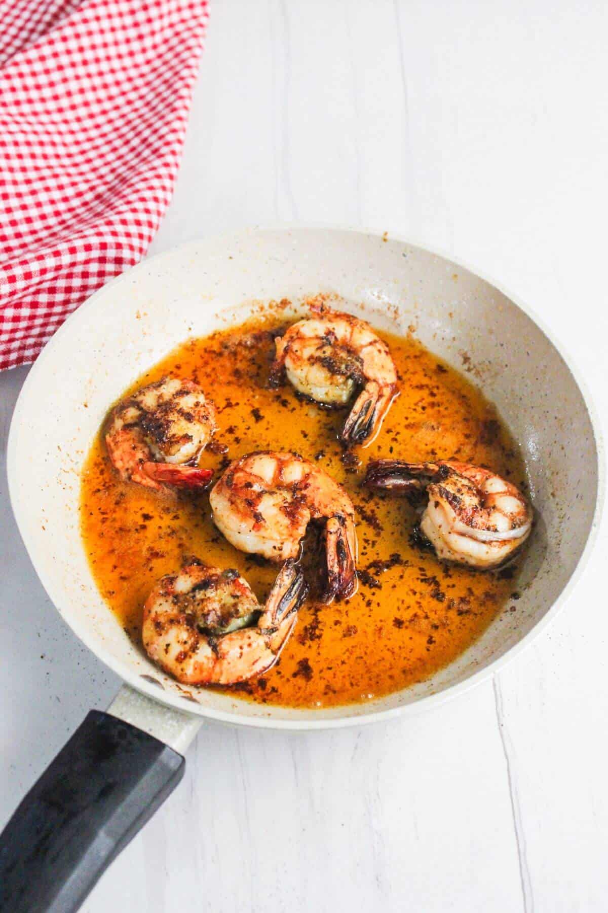 A white frying pan with four cooked shrimp in a seasoned orange sauce sits on a white surface; a red checkered cloth is nearby.