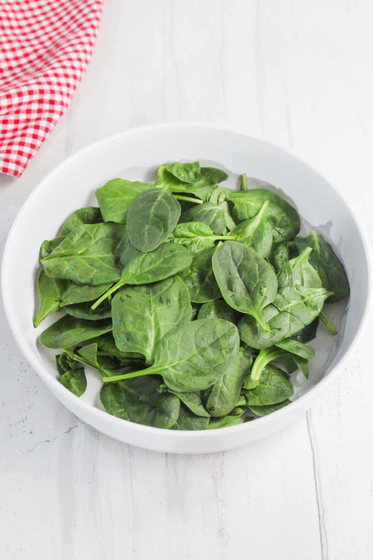 A white bowl filled with fresh spinach leaves sits on a white surface next to a red and white checkered cloth.