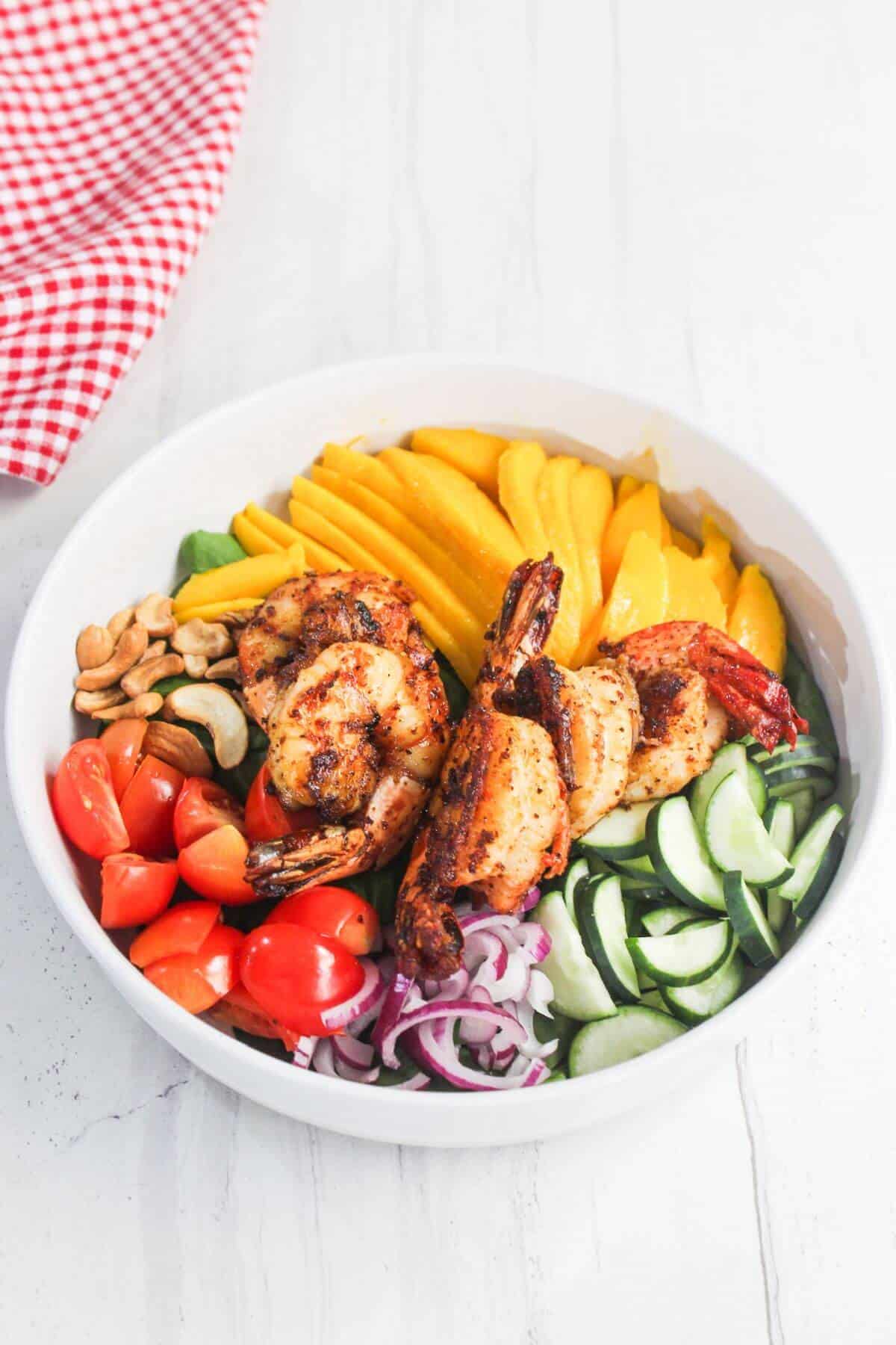 A white bowl filled with grilled shrimp, sliced mango, cherry tomatoes, red onions, cucumbers, cashews, and spinach on a white surface.