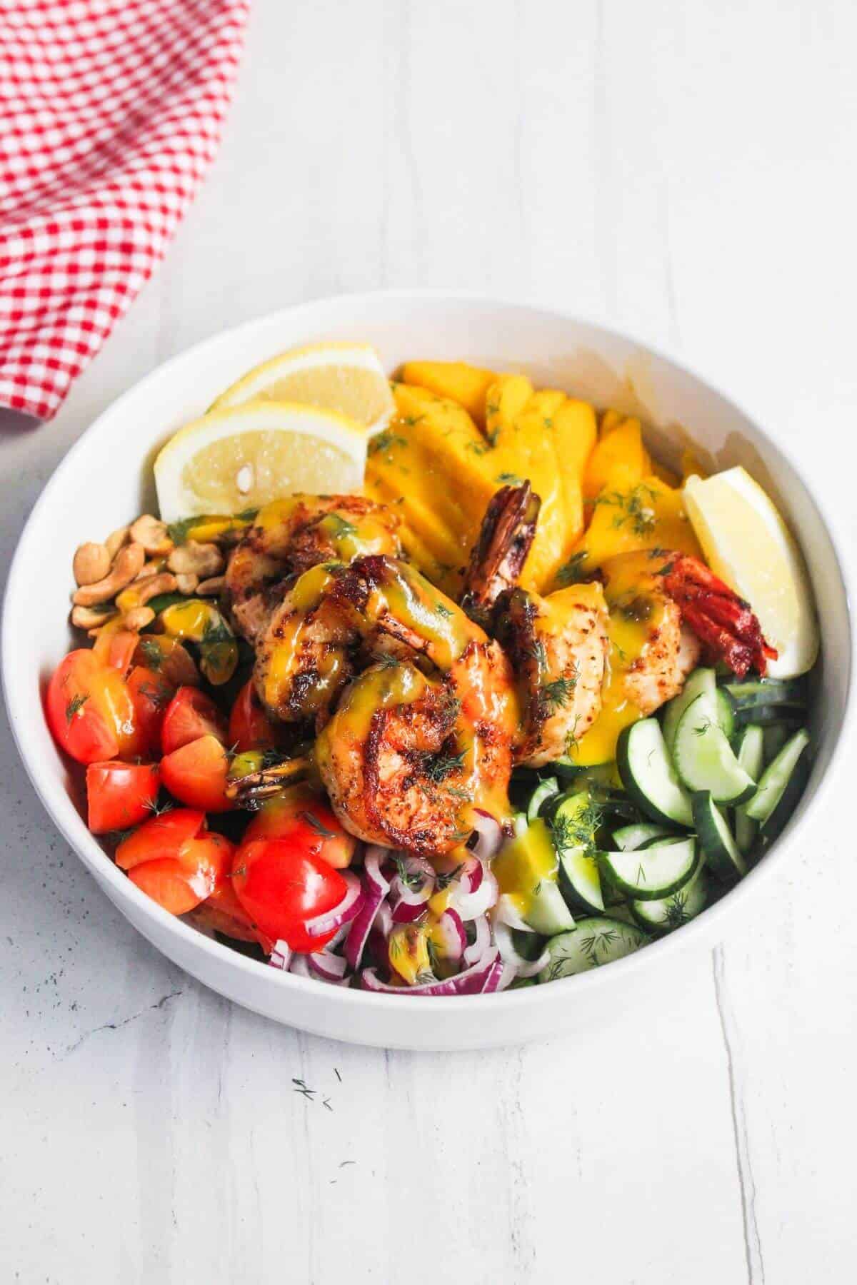 A bowl with grilled shrimp, cherry tomatoes, sliced cucumber, mango, red onion, lemon wedges, and peanuts, served on a white surface.