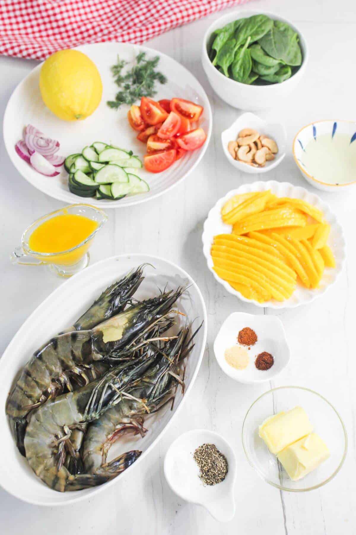 Assorted fresh ingredients on a white surface, including raw prawns, sliced cucumbers, tomatoes, spinach, mango, lemon, spices, butter, cashews, and a yellow sauce.